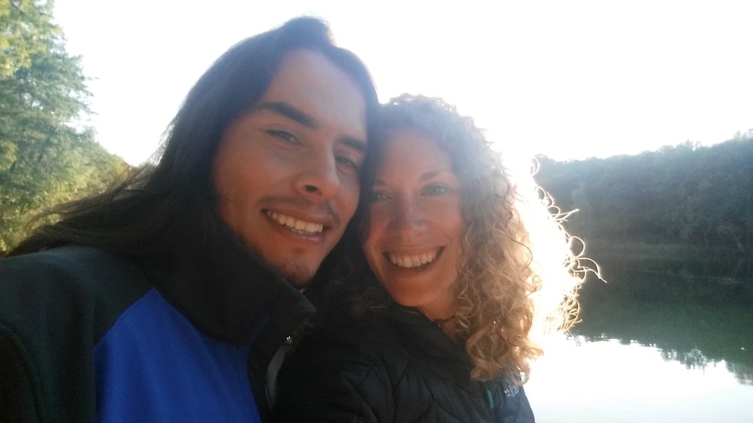 Picture shows Claire and Alfredo in a selfie, They are both smiling and in the background is a lake with trees visible on both sides of the water. The sky is bright white. 
