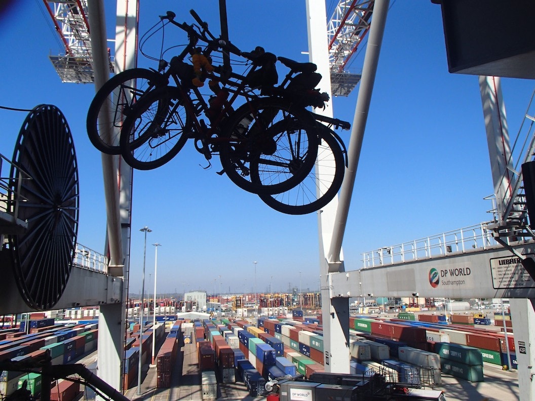 Picture shows two bikes being lifted over a large container ship. The sky is blue and the ship appears to large that you cant see where it ends. 