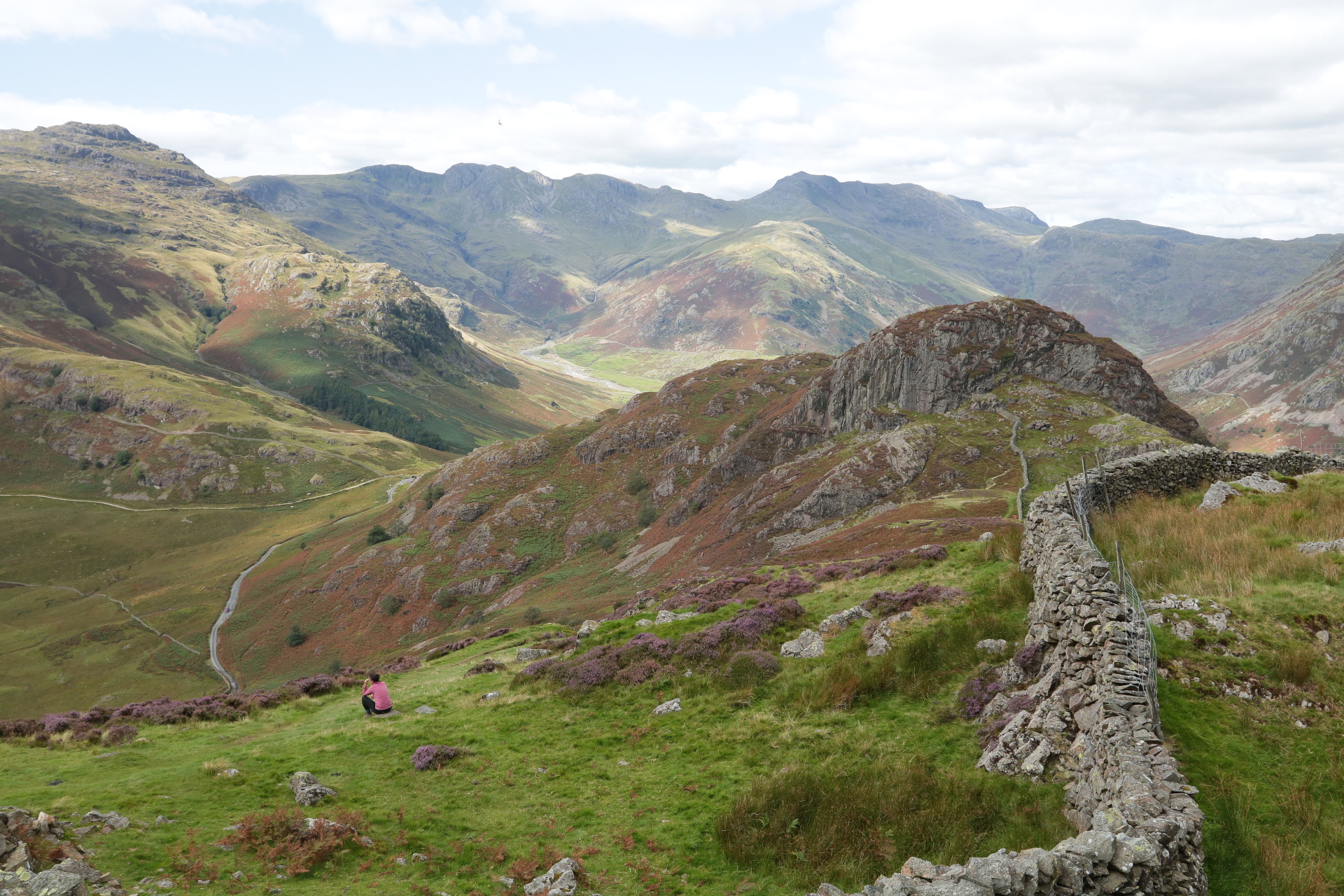 Image shows a wide view of the Lake District mountains. The grass is green with purple and red shrubs. Deep valleys are visible and the sun is shining in spots around the hills. You can see Sophia sitting on a rock. 