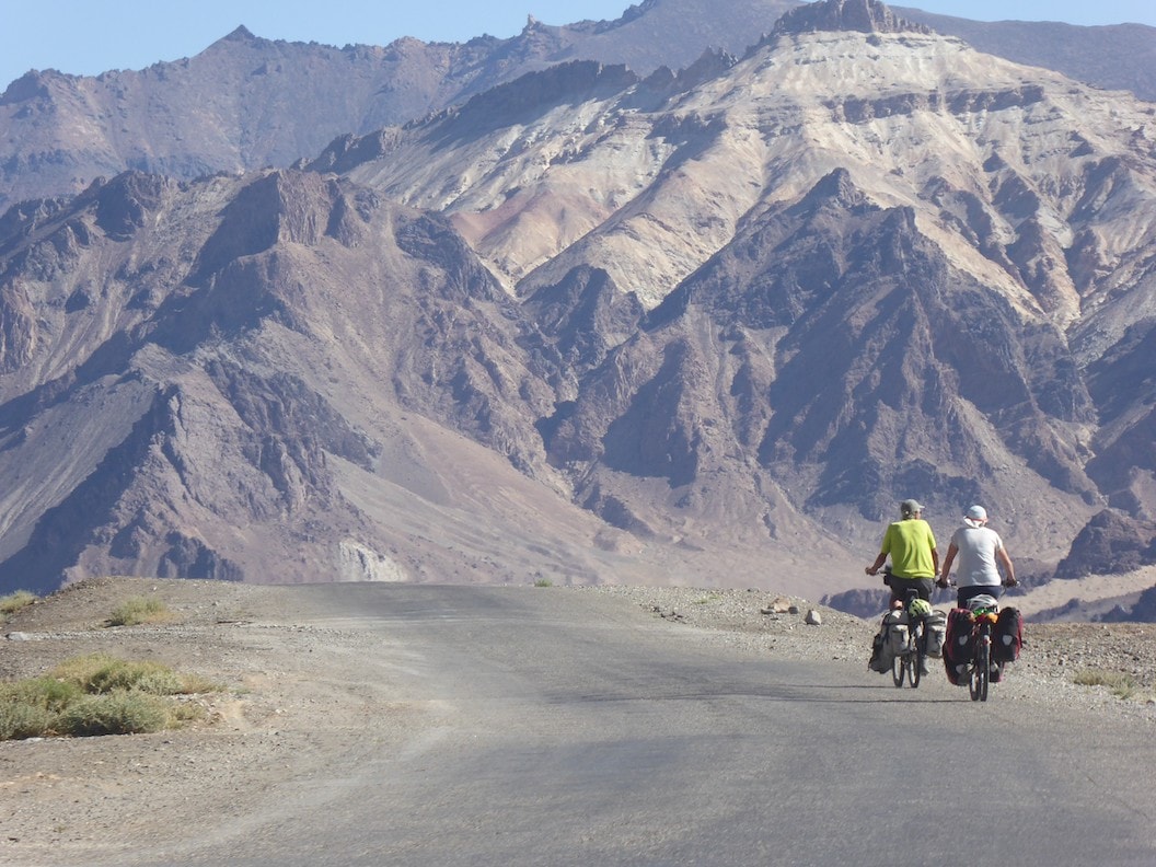 Picture shows Christine and Peter cycling along a dusty road in Tajikistan. They have their backs to the camera and in front of them is a vast mountainous landscape. 