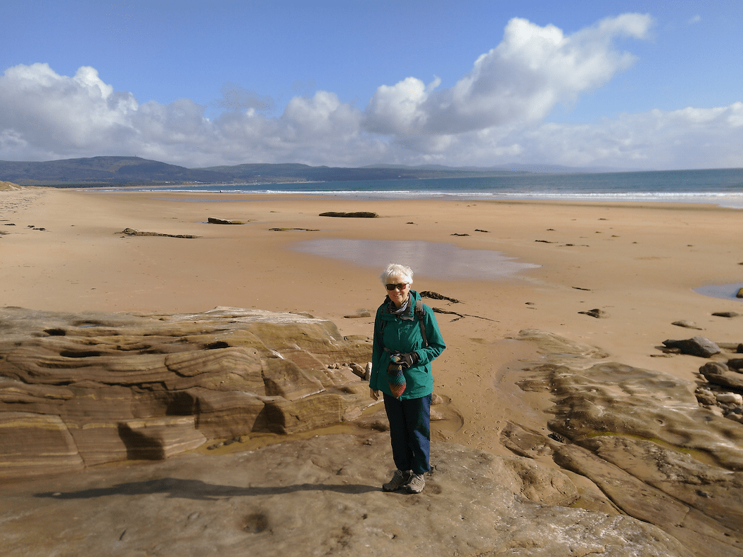 Picture shows Kate in a wide golden-sanded beach with the tide way out. There are smooth rocks surrounding her and clouds in the distance. 