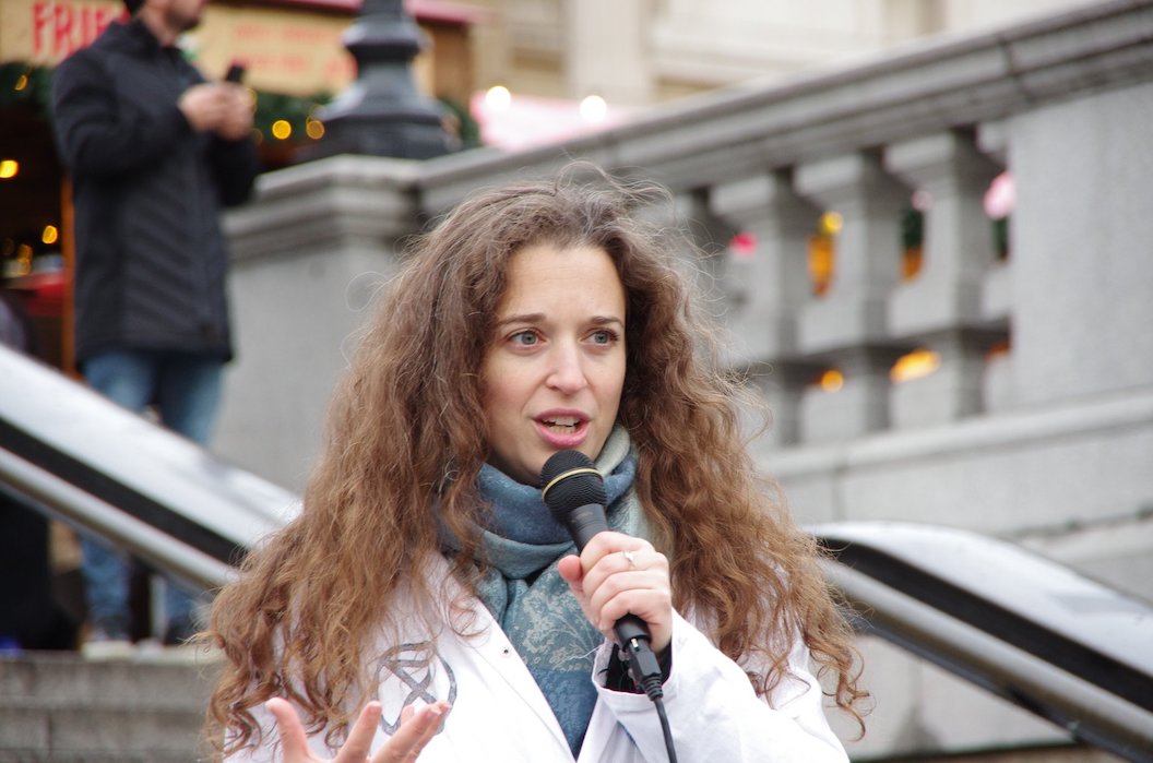 Dr Emily Grossman is speaking at an Extinction Rebellion protest, she is wearing a lab coat with the XR logo on it, and holding a microphone.