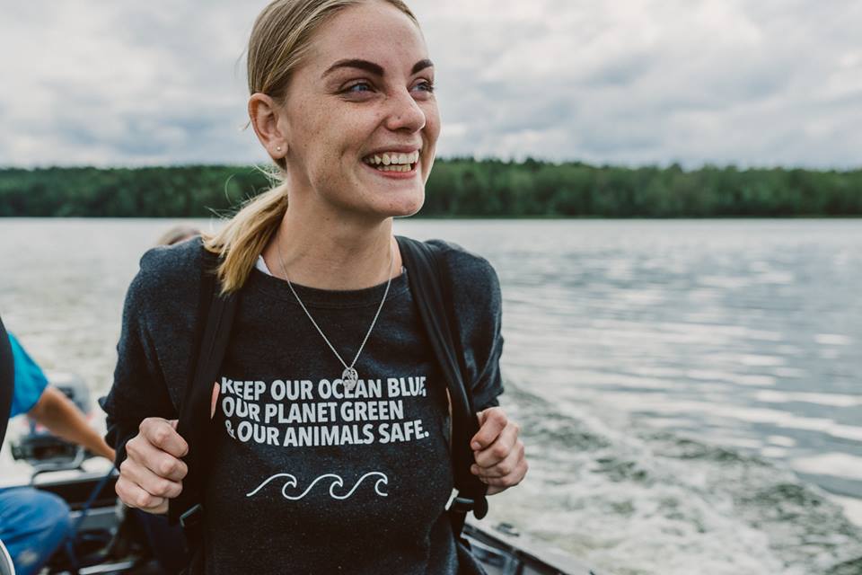 Evelina Evelina is smiling widely whilst being driven in a small boat. She is wearing a T-shirt that reads "Keep our oceans blue, our planet green and our animals safe". The are blurred trees in the background and an overcast sky.