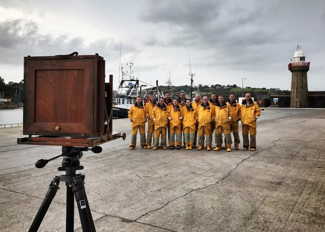 Picture shows an old fashion wooden standing camera taking a picture of a lifeboat team all wearing matching yellow boiler suits with yellow boots. They are standing on a concrete ramp into the sea, with boats in the background. The sky is pale grey.