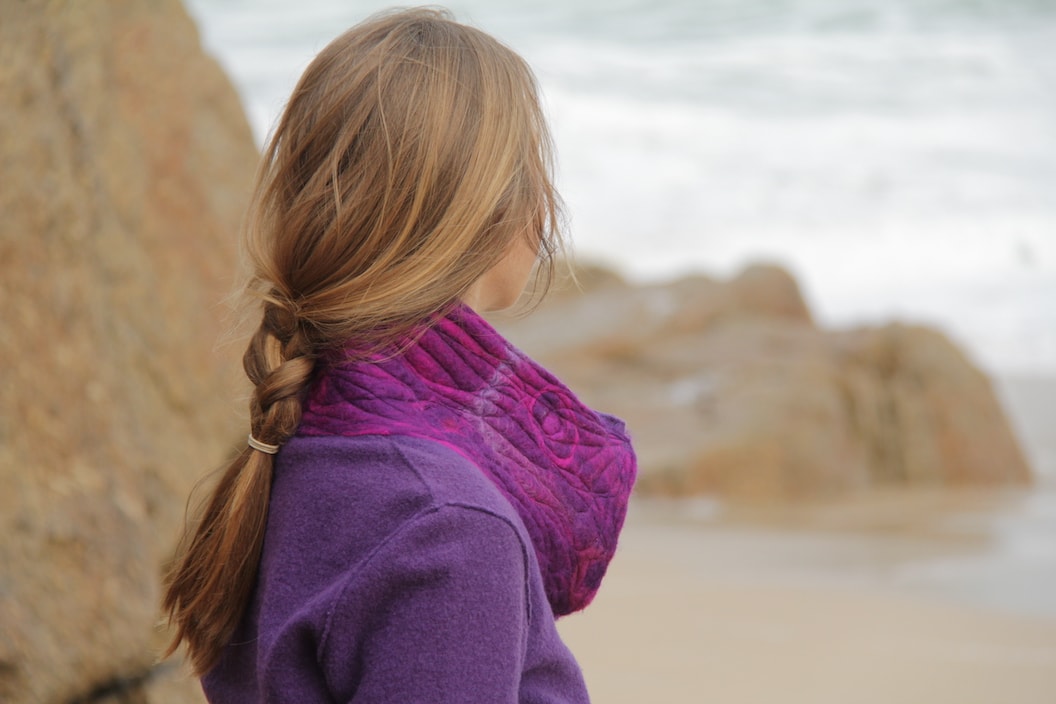 Picture shows a women wearing a purple jumper and a purple scarf. She is on the beach looking out to the sea and she has her back to the camera.