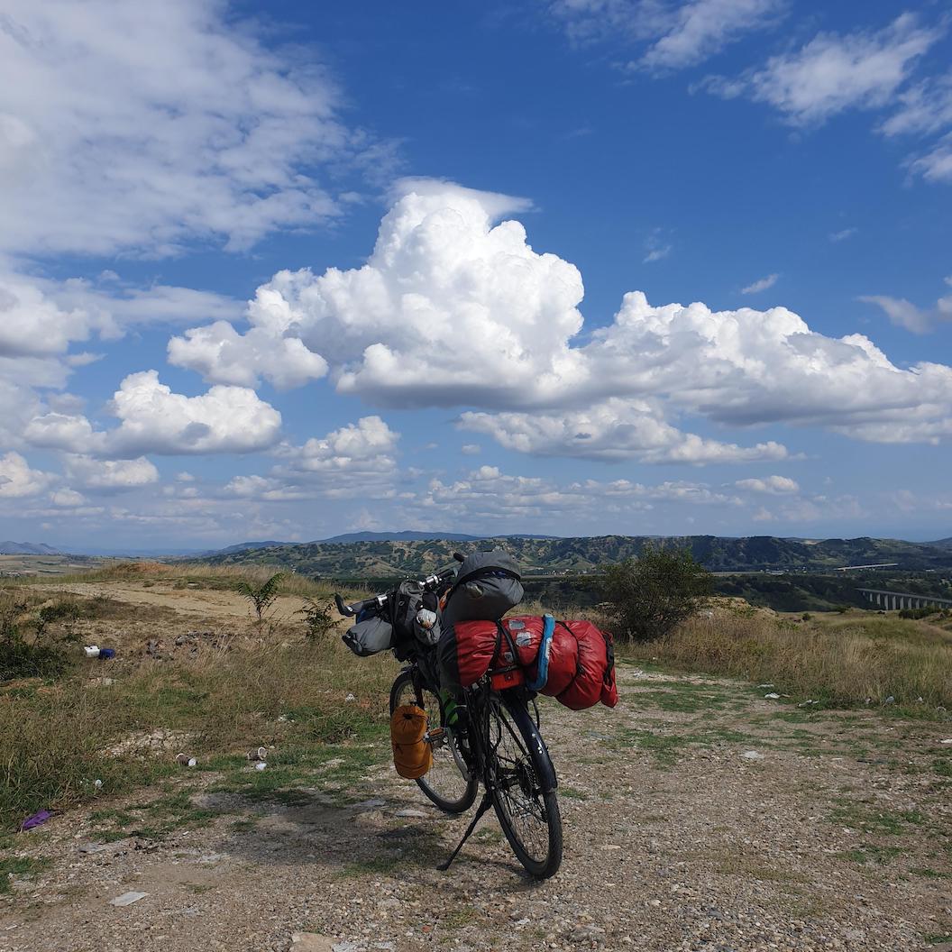 Picture shows a bicycle leaning on its stand packed with camping equipment. Behind it is a grassy and gravelly landscape with rolling hills in the background. 