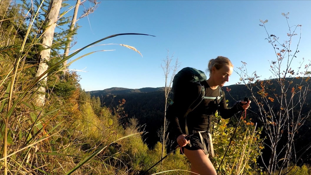 Picture shows Rosie hiking through tall grass on a sunny hillside. She is wearing activewear and carrying a large rucksack. There are tall hills in the background and the sky is bright blue. 