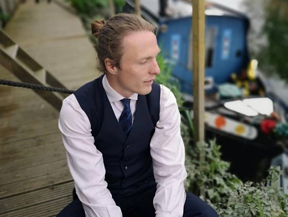 Picture shows Paul wearing a shirt, waistcoat and tie, sitting on decking next to his narrow boat on a canal. There are shrubs in the background and he is looking pensively away from the camera. 