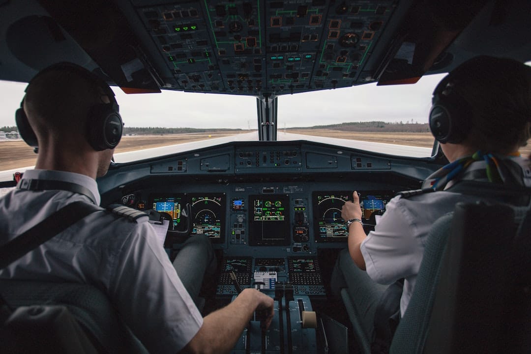 Photo shows two pilots from the back seated next to each other facing the cockpit of a plane. Ahead of them is a runway and it is a cloudy day. 