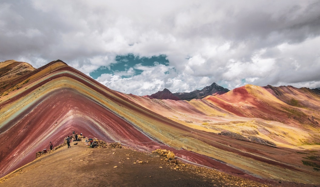 Picture shows the Rainbow Mountains. There is a mountain landscape, but each hill is made up of many horizontal slanting stripes of colour. the colours include deep burgundy, orange, sand, red and even eggshell blue. The clouds look moody and dramatic above. 