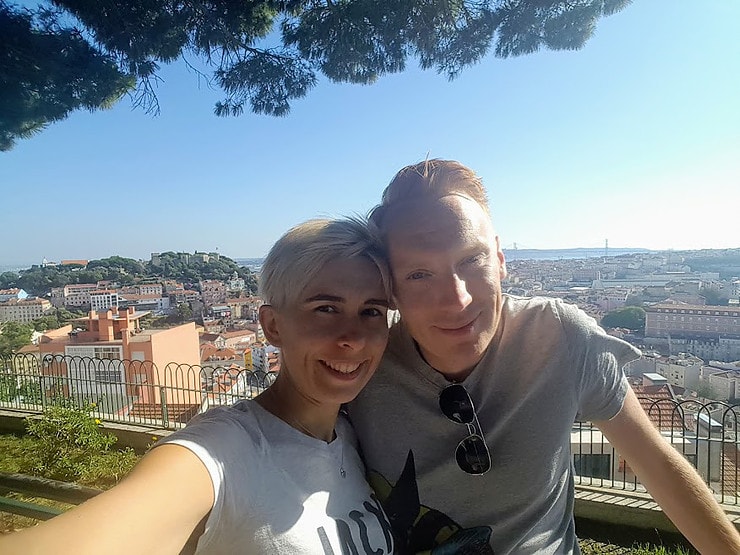 Marc and Rebecca take a selfie with a view of sunny Lisbon behind them. Many of the buildings have terracotta roofs and the sun is shining. 