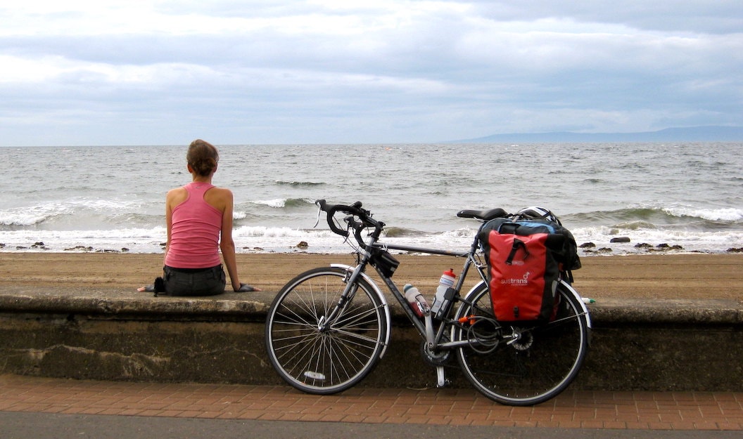 Anna sits with her back to the camera on a low wall separating the beach from the promenade. The beach is sandy and the waves and sky are moody and grey. Next to Anna resting against the wall is her bike. 