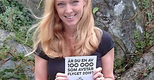 Picture of Maja Rosén holding a piece of paper with the flight free pledge on it in Swedish. She is sitting in front of a wall of large rocks covered in plants and moss. 