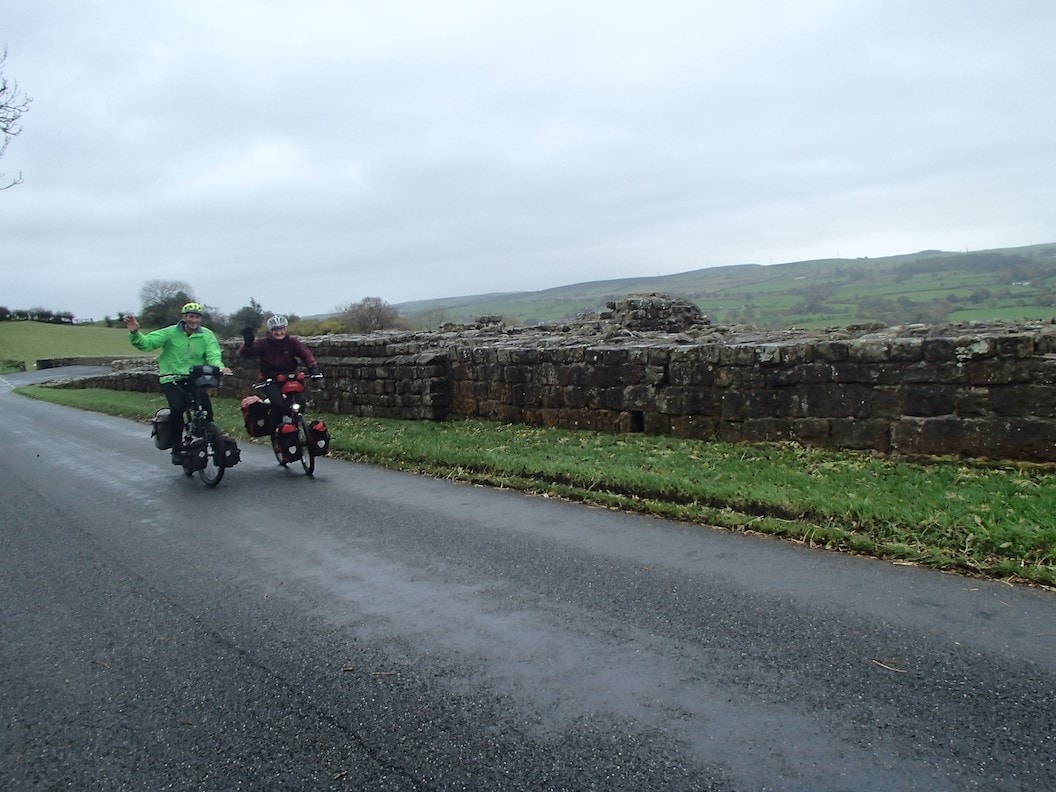 Picture shows Christine and Peter cycling next to Hadrians Wall. It is a gloomy day and the sky is overcast. There are fields in the background. 