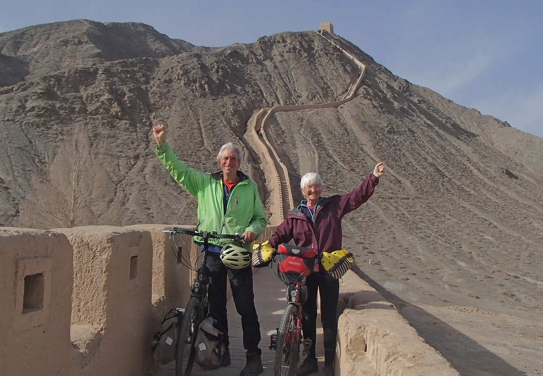 Picture shows Christine and Peter on a dusty and pale red part of the Great Wall of China. They are both holding their bikes with one hand and holding their fists in the air with the other. Peter is wearing a green coat and Christine is wearing a burgundy one. 